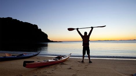 A man standing next to a kayak on the beach in Eden saluting the morning sun with his paddle
