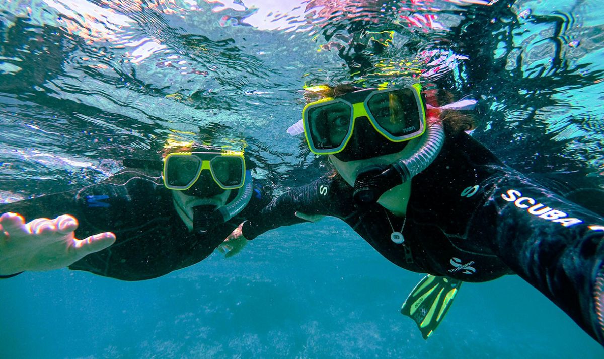 A close-up of two people with snorkelling gear of looking at the camera while floating just under the surface of the water